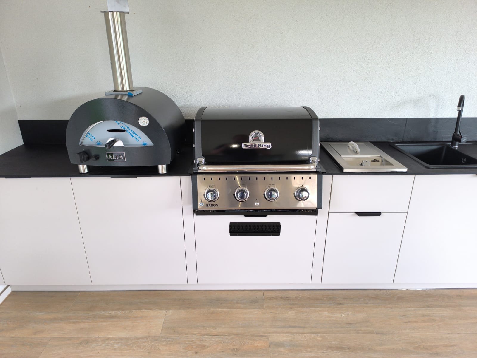 Zewnętrzna kuchnia z piecem do pizzy Alfa i grillem Broil King Baron na białych szafkach z czarnym blatem, widok z przodu, jasne wnętrze, drewniana podłoga.