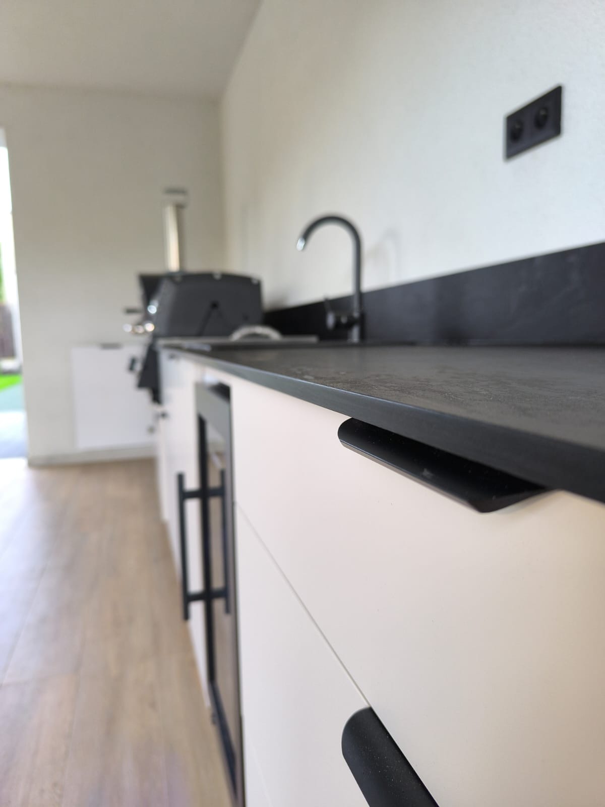 Nowoczesna, zewnętrzna kuchnia z blatem z ciemnego kamienia i białymi szafkami z czarnymi uchwytami. W tle grill i zlew.