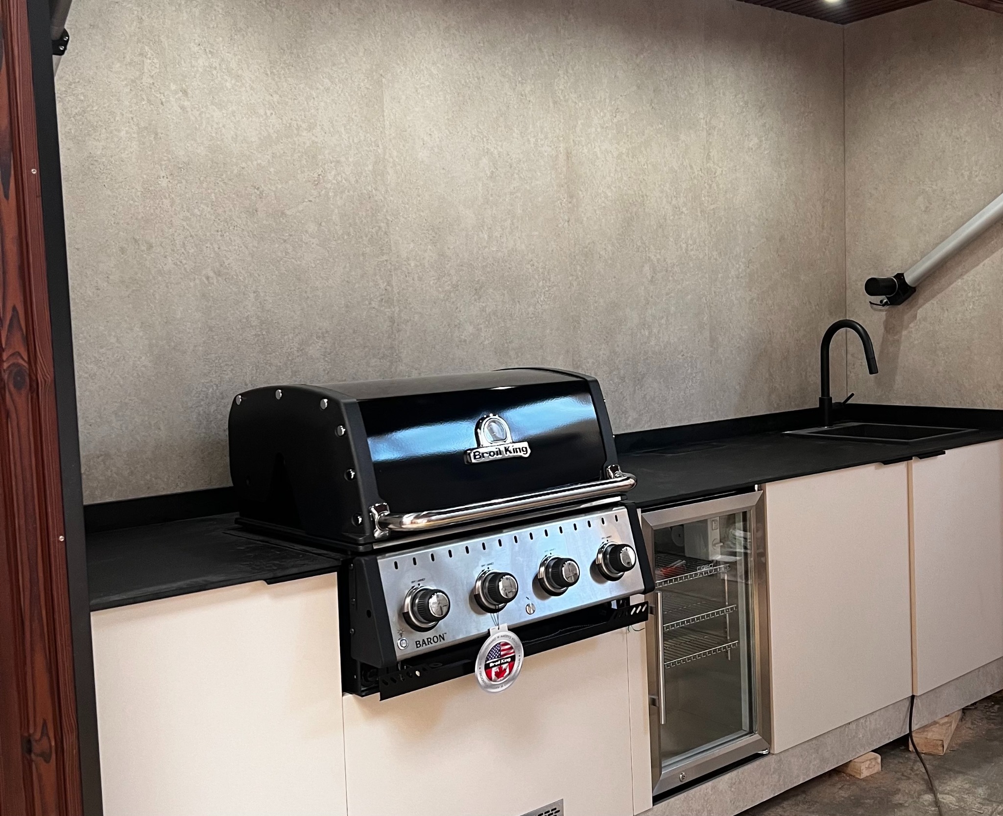 Zadaszona kuchnia zewnętrzna z grillem Broil King Baron, lodówką podblatową i czarnym blatem. Nowoczesny design, jasne szafki, szara ściana. Ujęcie z bliska.