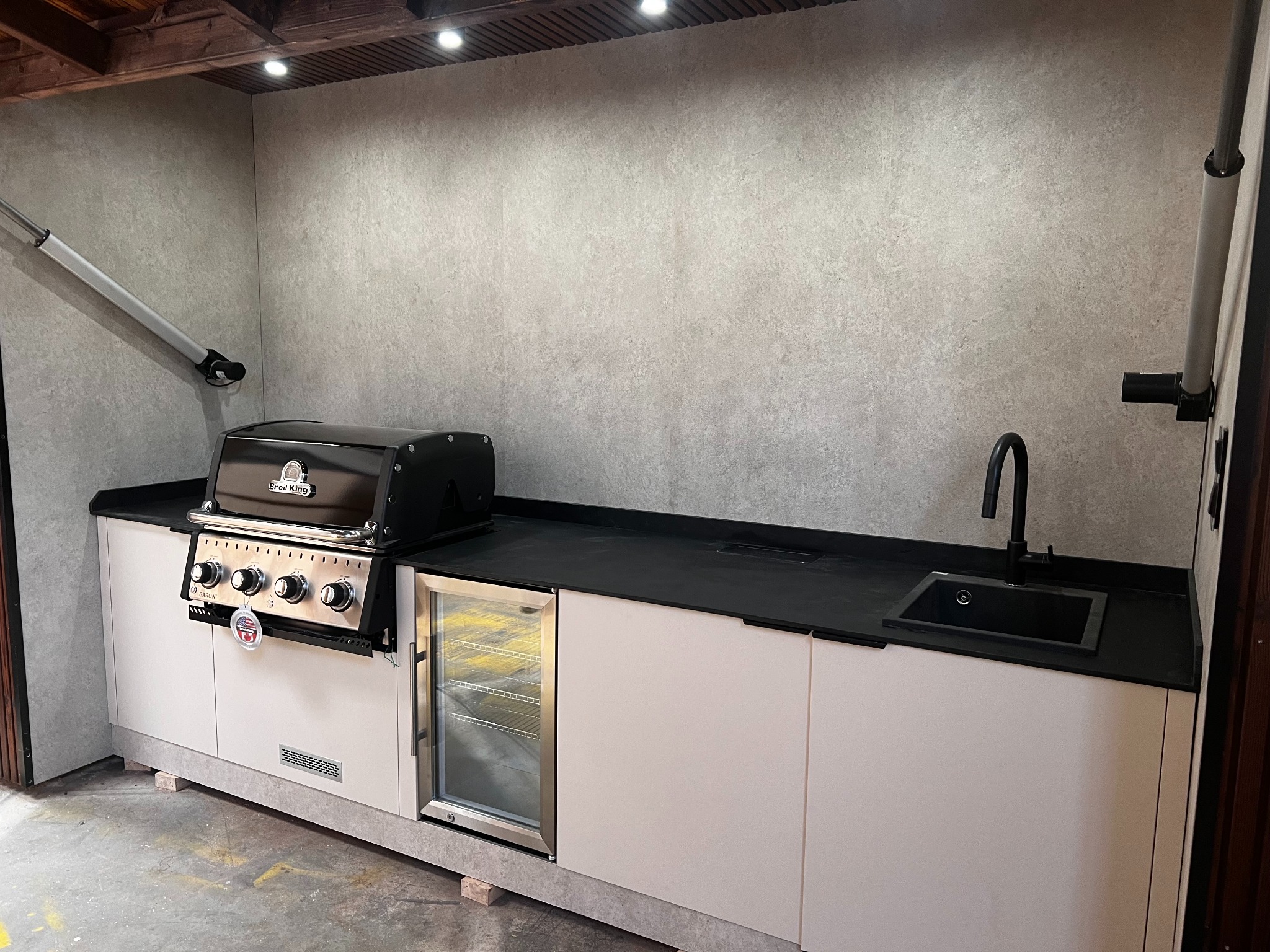 Zadaszona kuchnia zewnętrzna z grillem Broil King, lodówką na napoje i zlewem z czarną armaturą, wykończona w szarościach i bieli, oświetlona punktowo.