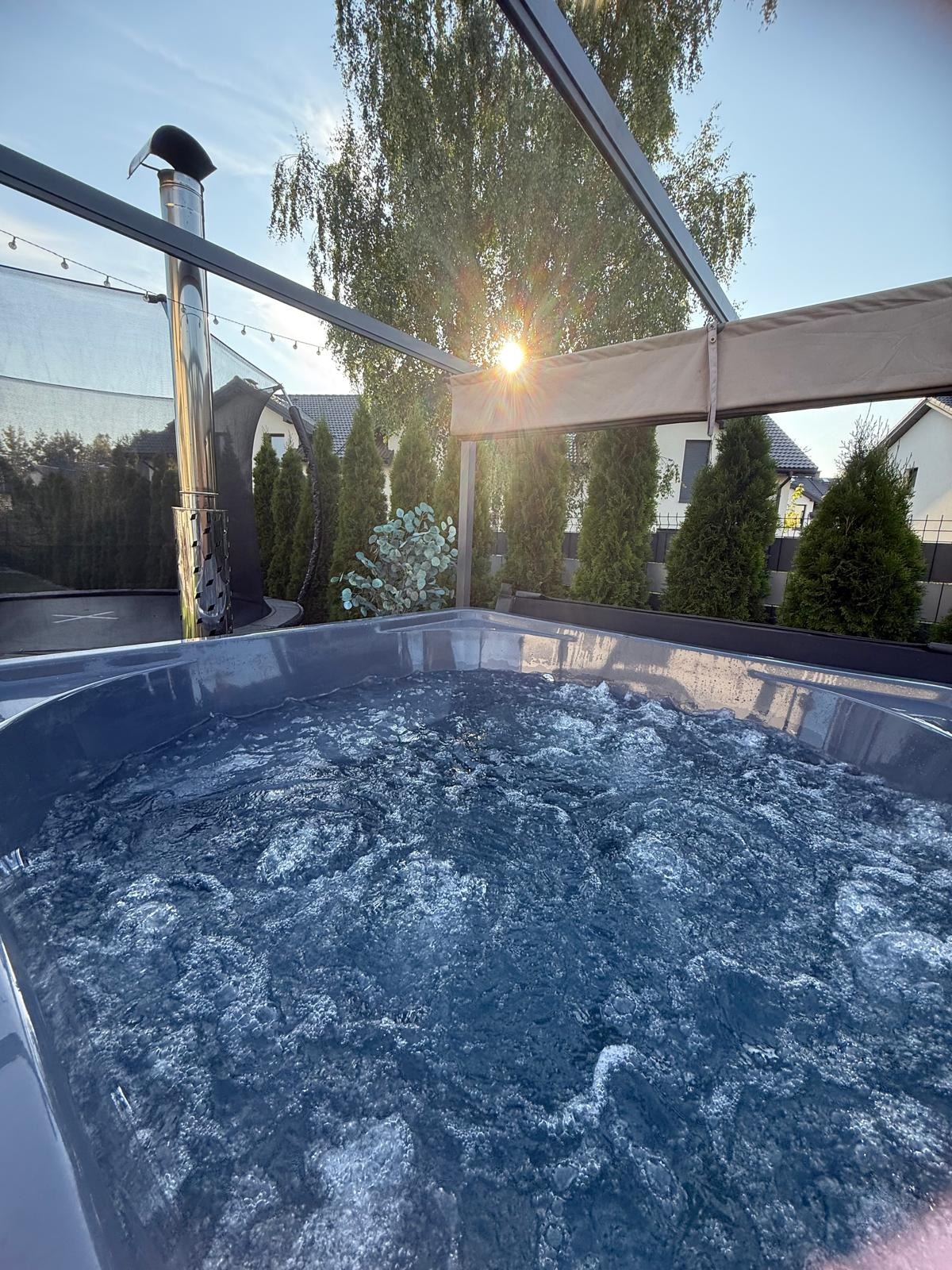 Nowoczesna wanna spa z hydromasażem na zewnątrz, pod metalową pergolą. W tle zielone tuje i domy. Słońce przebija przez drzewo.