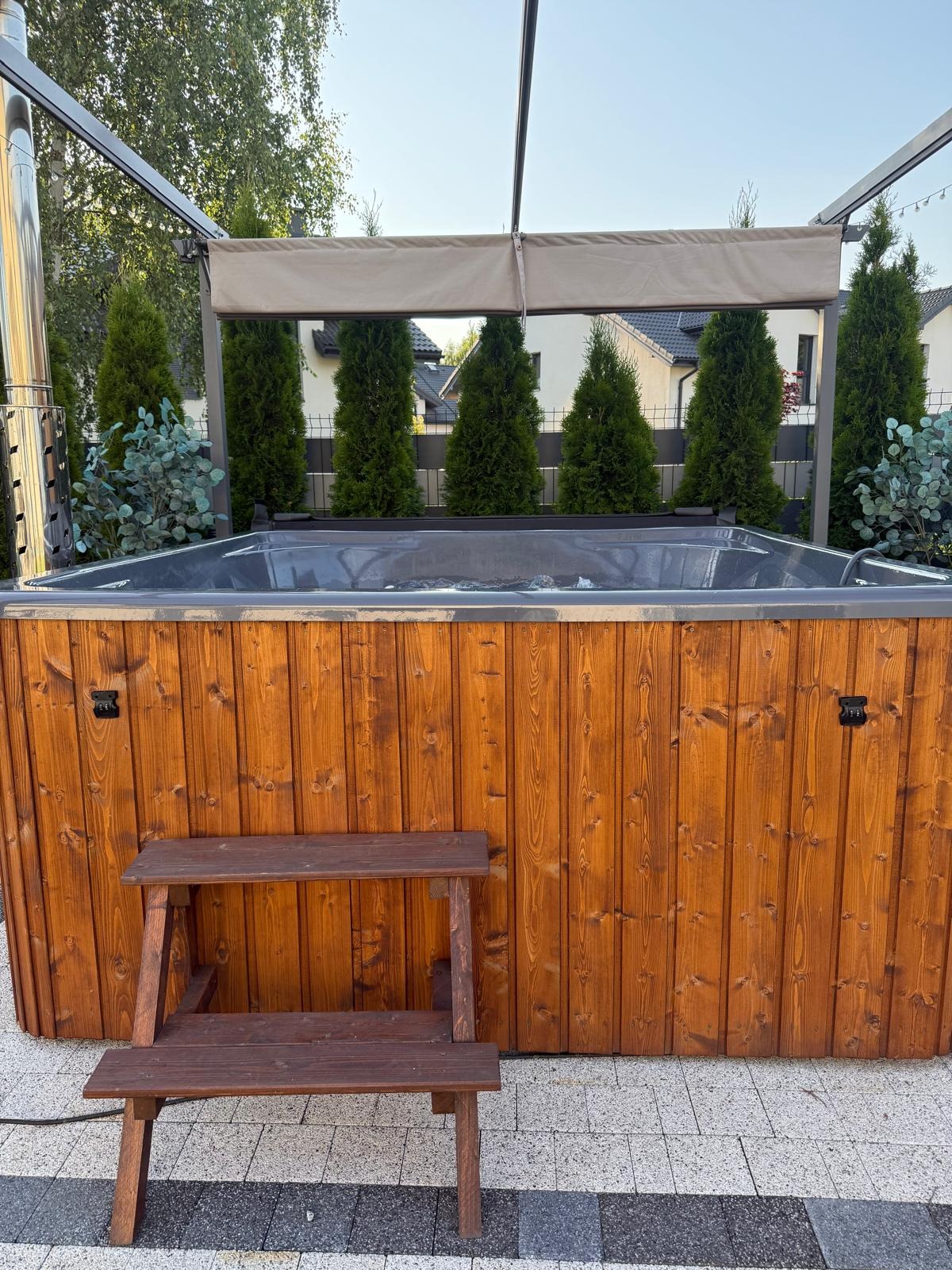 Drewniana sauna zewnętrzna z zadaszeniem i schodkami, otoczona tujami, na tle domu jednorodzinnego w słoneczny dzień. Widok z przodu, detale wykończenia.