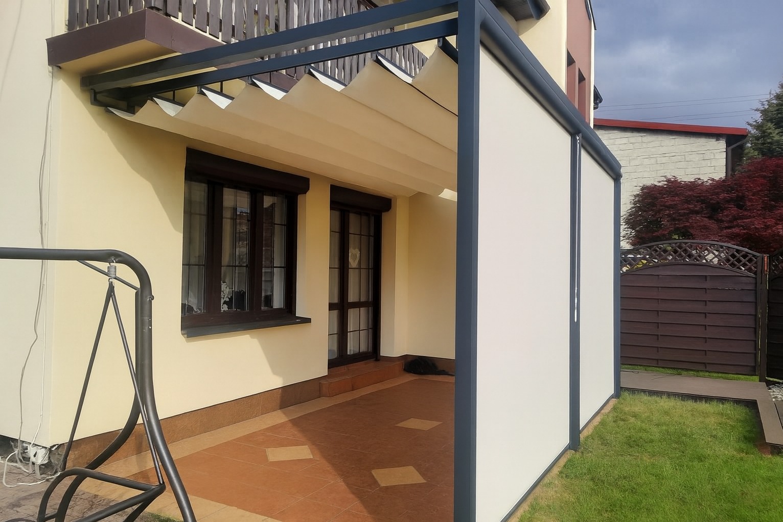 Nowoczesna pergola z przesuwnymi panelami bocznymi, przylegająca do budynku. Kremowa elewacja, brązowe okna i drzwi. Widoczny fragment huśtawki ogrodowej.