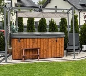 Zadaszona, metalowa pergola w kolorze grafitowym, osłaniająca drewnianą balię z hydromasażem, otoczoną kostką brukową i zielonym trawnikiem. W tle dom i tuje.