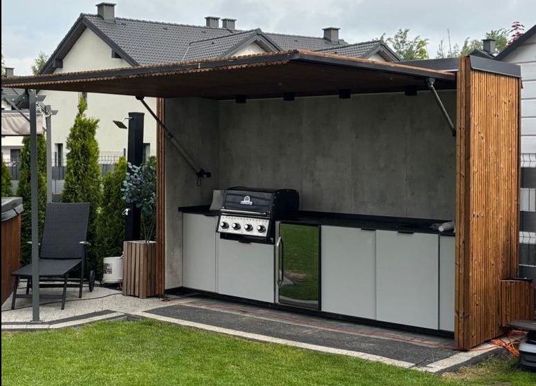 Zadaszona kuchnia letnia z grillem i szafkami, wykończona drewnem i betonem, z widocznym fragmentem ogrodu i domu w tle. Elementy drewniane pionowe.