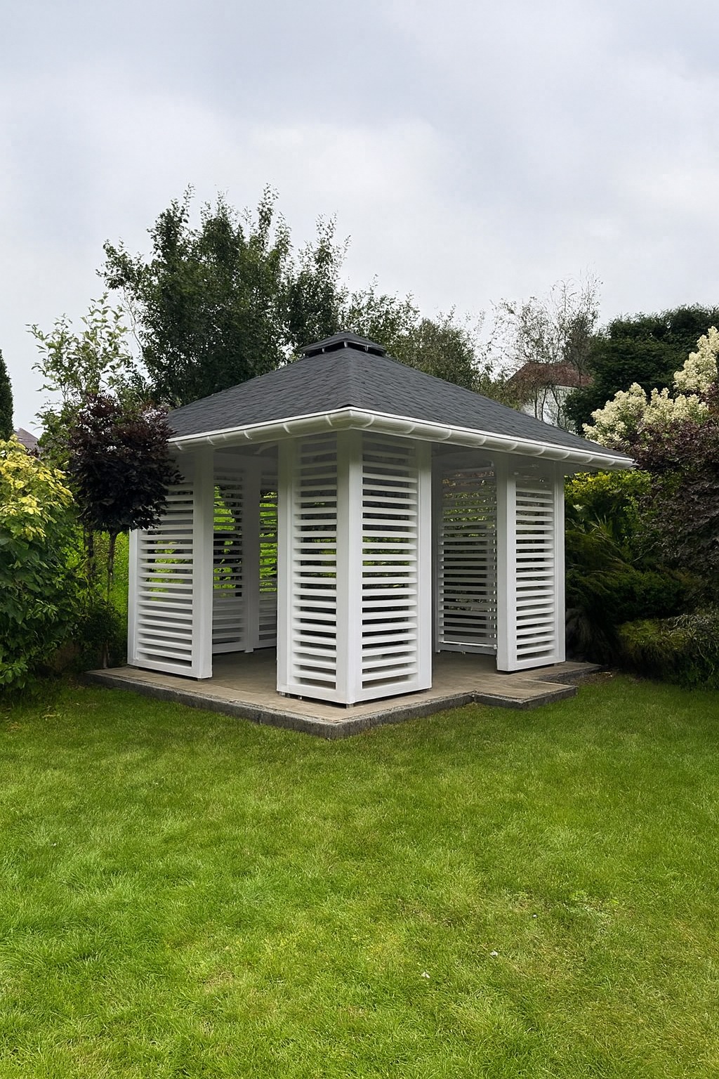 Nowoczesna, biała pergola z poziomymi panelami w ogrodzie. Czarny, spadzisty dach, szara kostka brukowa, zielony trawnik i drzewa w tle. Minimalistyczny design, naturalne otoczenie.