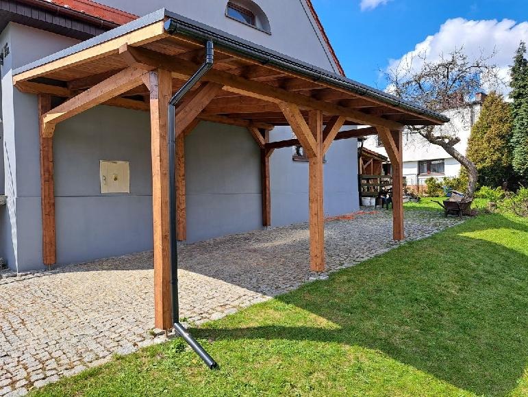 Drewniana pergola przy domu z szarym tynkiem, widoczny system odprowadzania wody. Konstrukcja oparta na czterech słupach, zadaszenie z desek, kostka brukowa na podłożu.