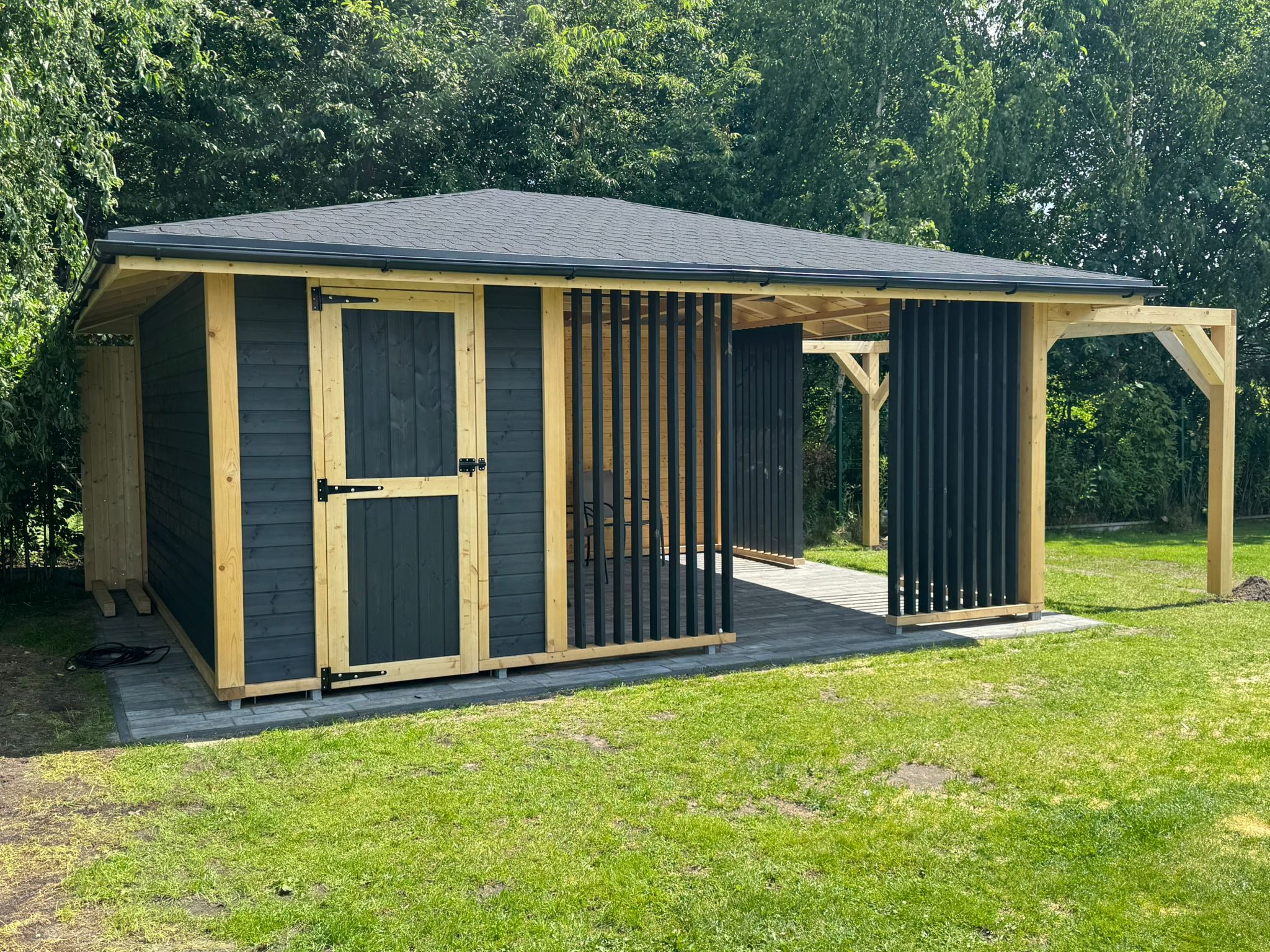 Nowoczesna, drewniana altana z czarnymi elementami, wykończona gontem bitumicznym, na tle zielonego trawnika i drzew. Częściowo otwarta konstrukcja.