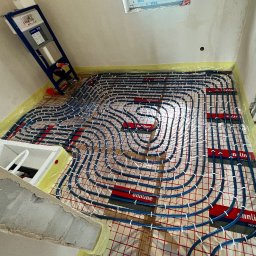 HYDROPIK Paweł Nowak - Niebieskie rury systemu ogrzewania podłogowego ułożone na czerwonej siatce montażowej, widok z góry, w tle częściowo widoczna instalacja sanitarna podtynkowa.