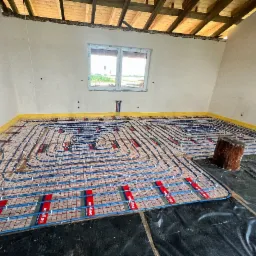 Niedokończona instalacja ogrzewania podłogowego z niebieskimi rurkami na folii z czerwonymi paskami w surowym wnętrzu poddasza z drewnianą konstrukcją dachu i oknem.