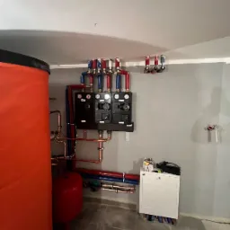 Pomieszczenie techniczne z dużym, pomarańczowym zbiornikiem, dwoma czarnymi modułami sterującymi, instalacją z rur miedzianych i niebiesko-czerwonych, oraz białą szafką z narzędziami na górze.
