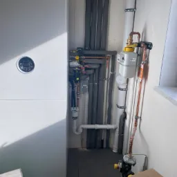 Biała pompa ciepła Bosch z widocznymi rurami miedzianymi, szarymi izolowanymi przewodami i licznikiem, zamontowana przy szarej ścianie. Fragment kartonów na podłodze.