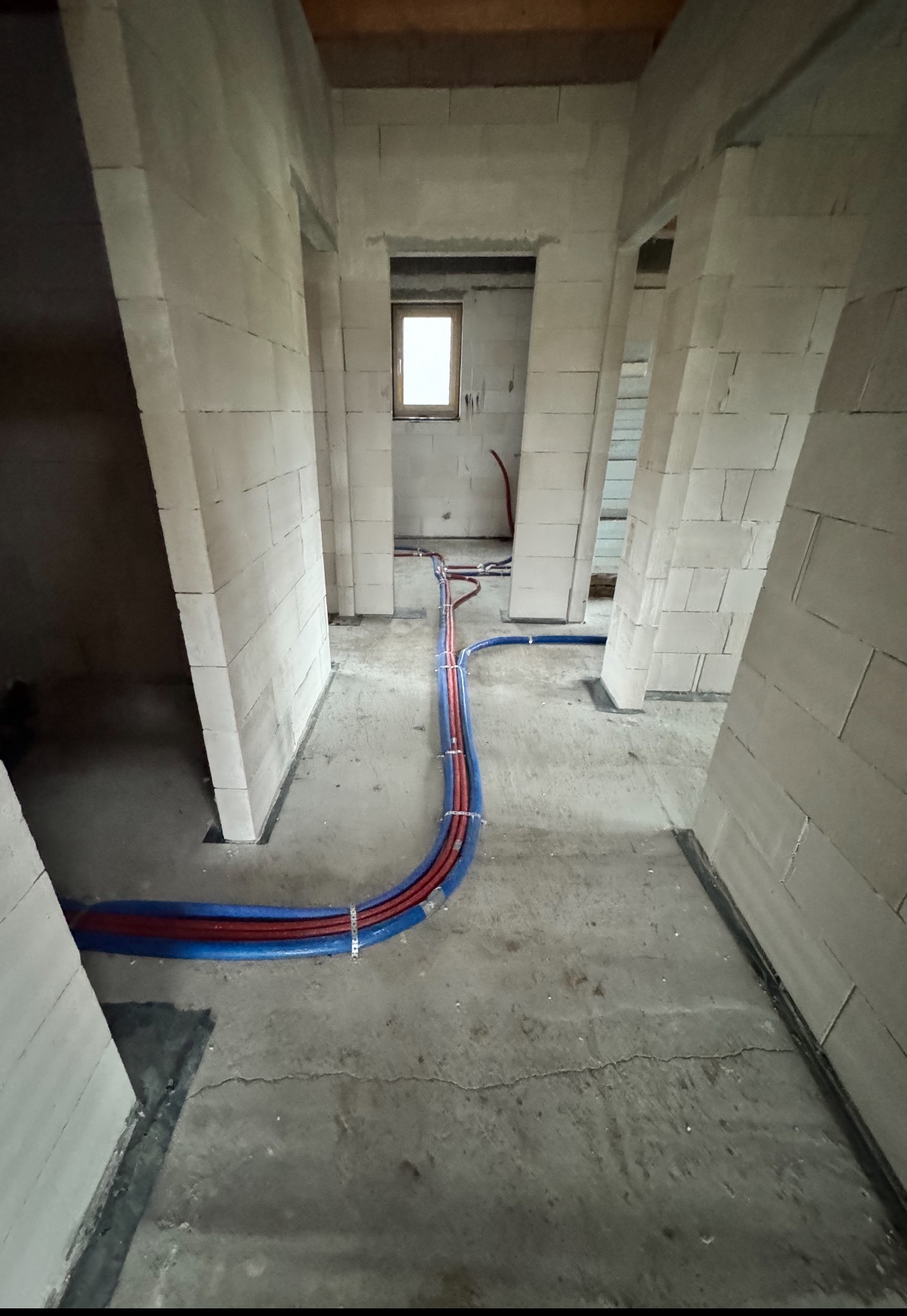 Instalacja ogrzewania podłogowego w surowym stanie: rury grzewcze czerwono-niebieskie przymocowane do podłoża klamrami, widok na wnętrze budynku z bloczków.