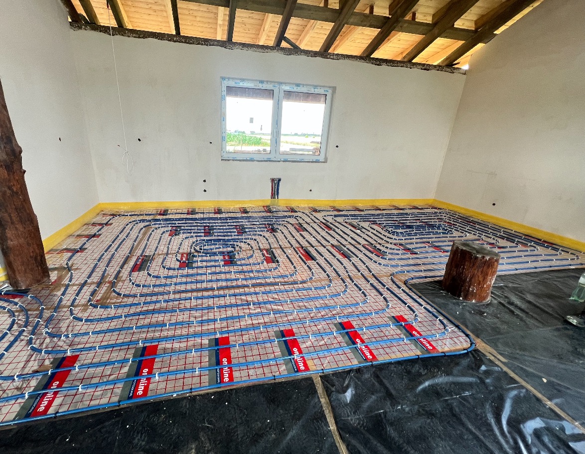 Niedokończona instalacja ogrzewania podłogowego z niebieskimi rurkami na folii z czerwonymi paskami w surowym wnętrzu poddasza z drewnianą konstrukcją dachu i oknem.