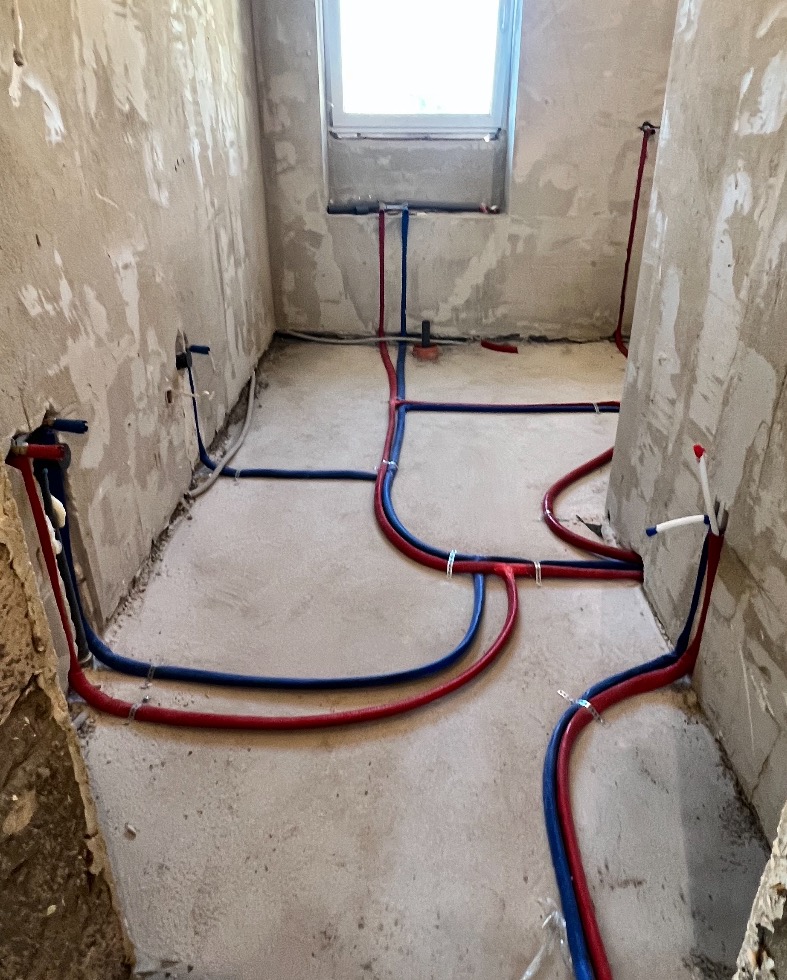 Surowe wnętrze łazienki w trakcie instalacji wodnej z widocznymi rurami w kolorach czerwonym i niebieskim, mocowanymi do surowych ścian i podłogi za pomocą metalowych uchwytów.