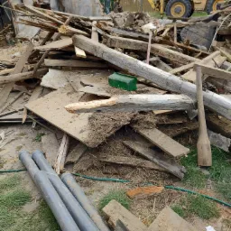 Stos gruzu i drewna po rozbiórce budynku, z widocznym żółtym traktorem w tle oraz fragmentami zniszczonej konstrukcji budynku.