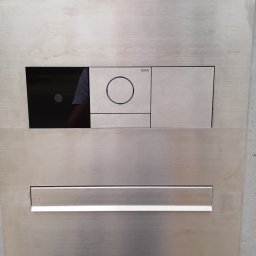 Łukasz Wojtunik Automatyka budynkowa - Szczegółowy widok panelu sterowania Gira ze stali nierdzewnej, wbudowanego w surowy betonowy mur, z widocznym odbiciem osoby w czarnym panelu dotykowym.