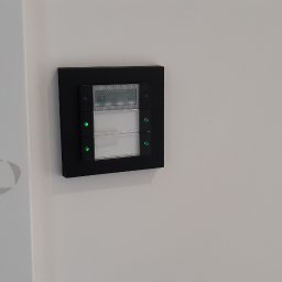 Łukasz Wojtunik Automatyka budynkowa - Panel sterowania inteligentnego domu z wyświetlaczem temperatury i ikoną chmury, zamontowany na białej ścianie obok drzwi z logo Rehau Rauvisio.