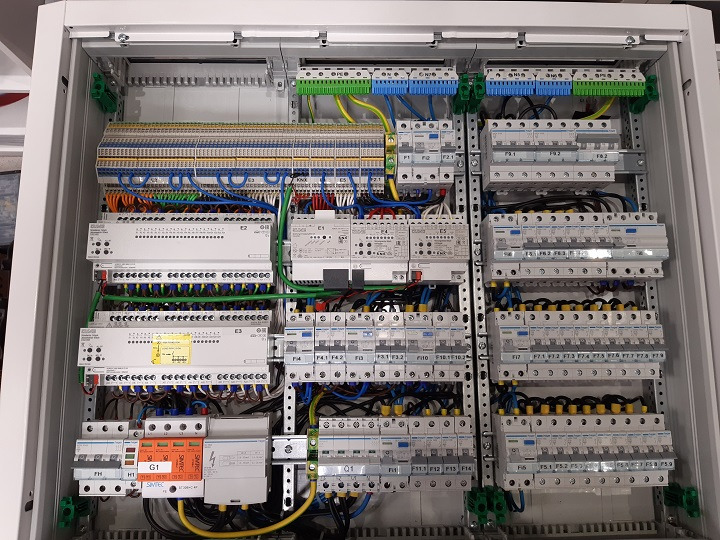 Szczegółowe ujęcie wnętrza rozdzielnicy elektrycznej z licznymi bezpiecznikami, modułami sterującymi i okablowaniem, widoczne oznaczenia na elementach i listwach zaciskowych.