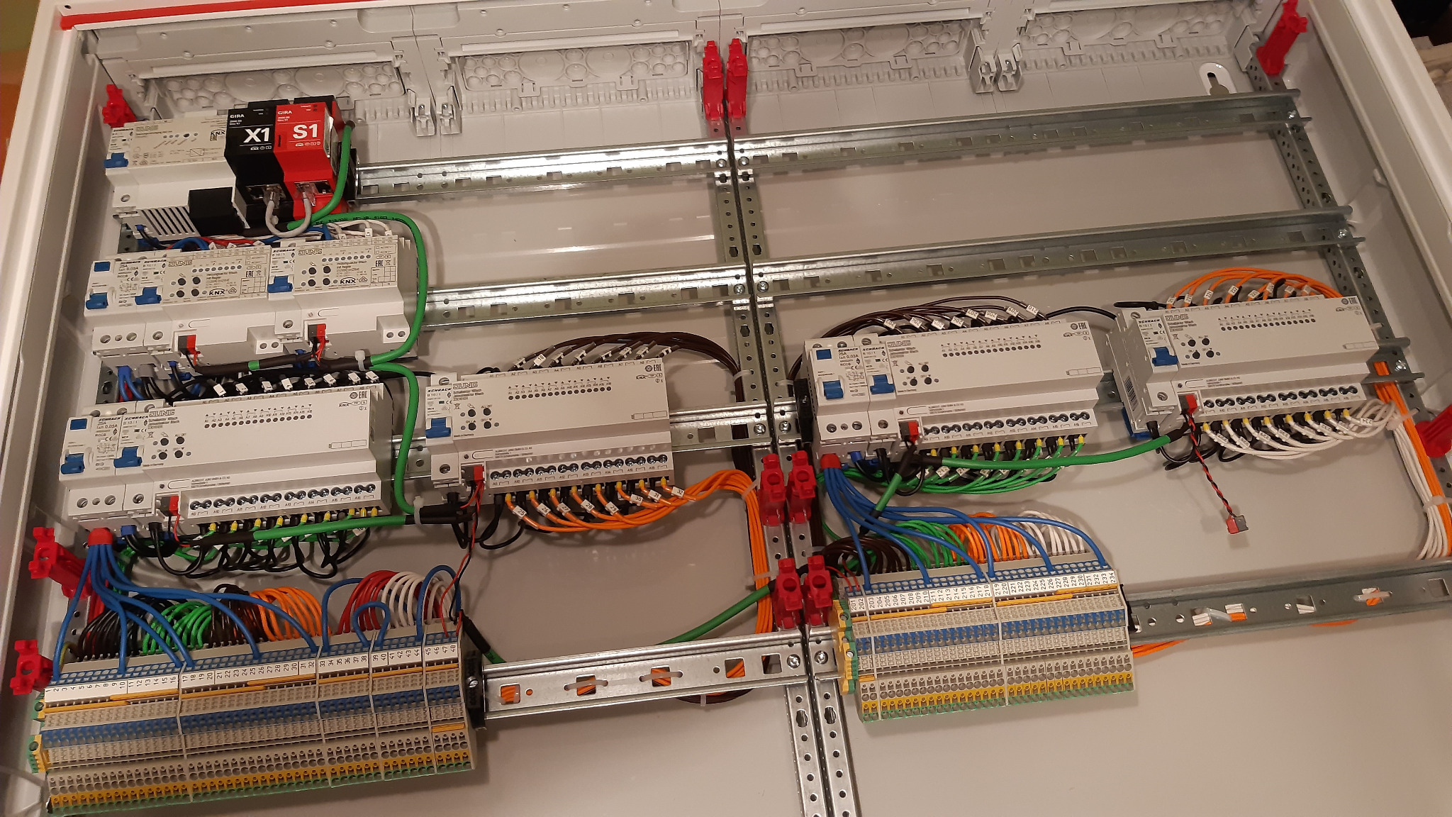 Otwarta skrzynka rozdzielcza z zamontowanymi modułami sterującymi KNX, licznymi przewodami w różnych kolorach i listwami zaciskowymi, widoczne metalowe szyny montażowe i elementy mocujące.