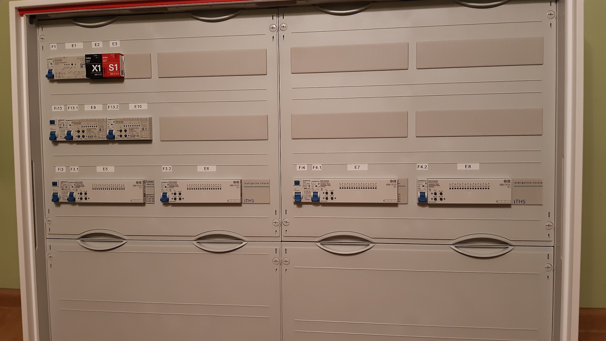 Szara rozdzielnica elektryczna z zamontowanymi modułami KNX, oznaczona etykietami F1, E1, E2, E3, Fi13, F13.1, E9, F13.2, E10, Fi3, F3.1, E5, F3.2, E6, F4, F4.1, E7, F4.2, E8, z widocznymi...