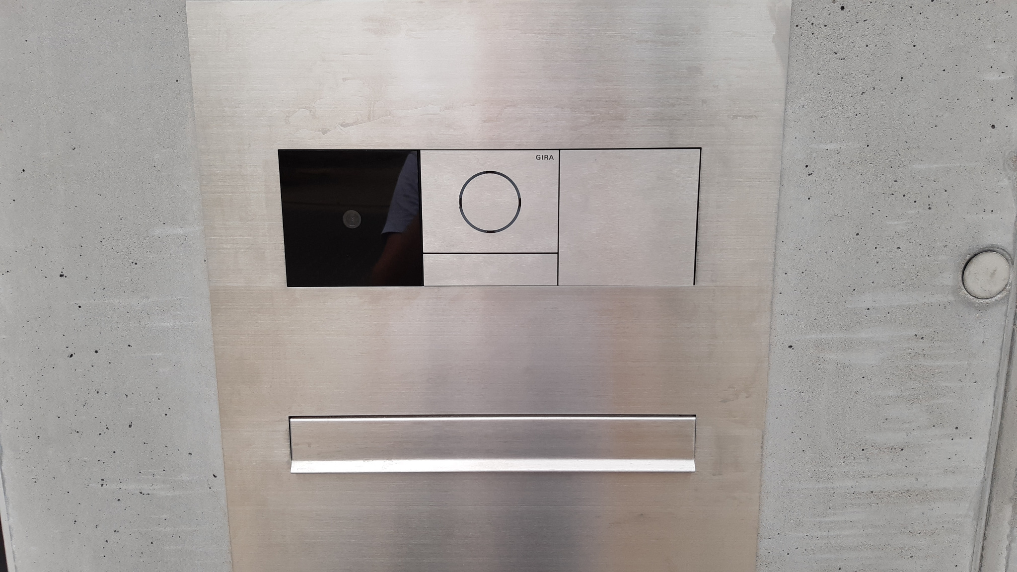 Szczegółowy widok panelu sterowania Gira ze stali nierdzewnej, wbudowanego w surowy betonowy mur, z widocznym odbiciem osoby w czarnym panelu dotykowym.