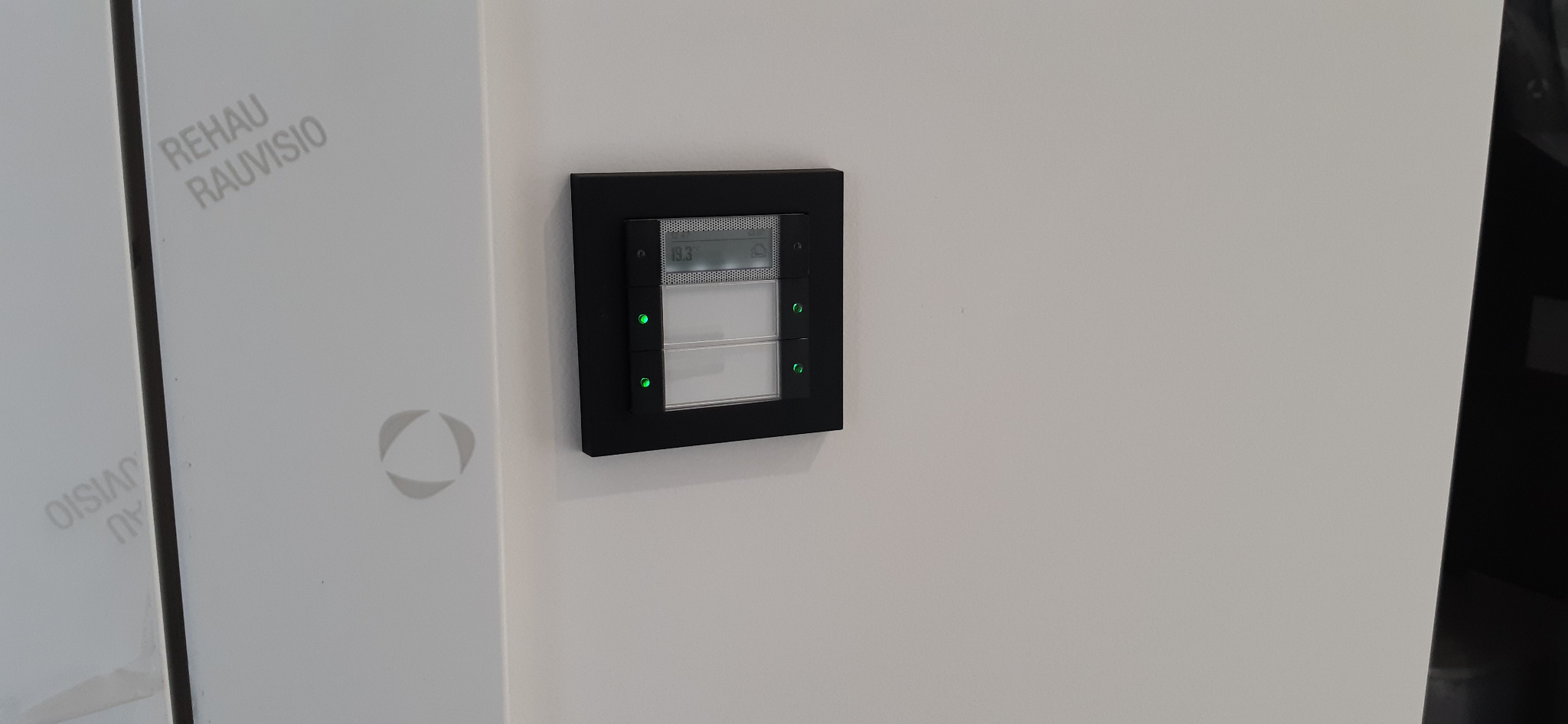 Panel sterowania inteligentnego domu z wyświetlaczem temperatury i ikoną chmury, zamontowany na białej ścianie obok drzwi z logo Rehau Rauvisio.