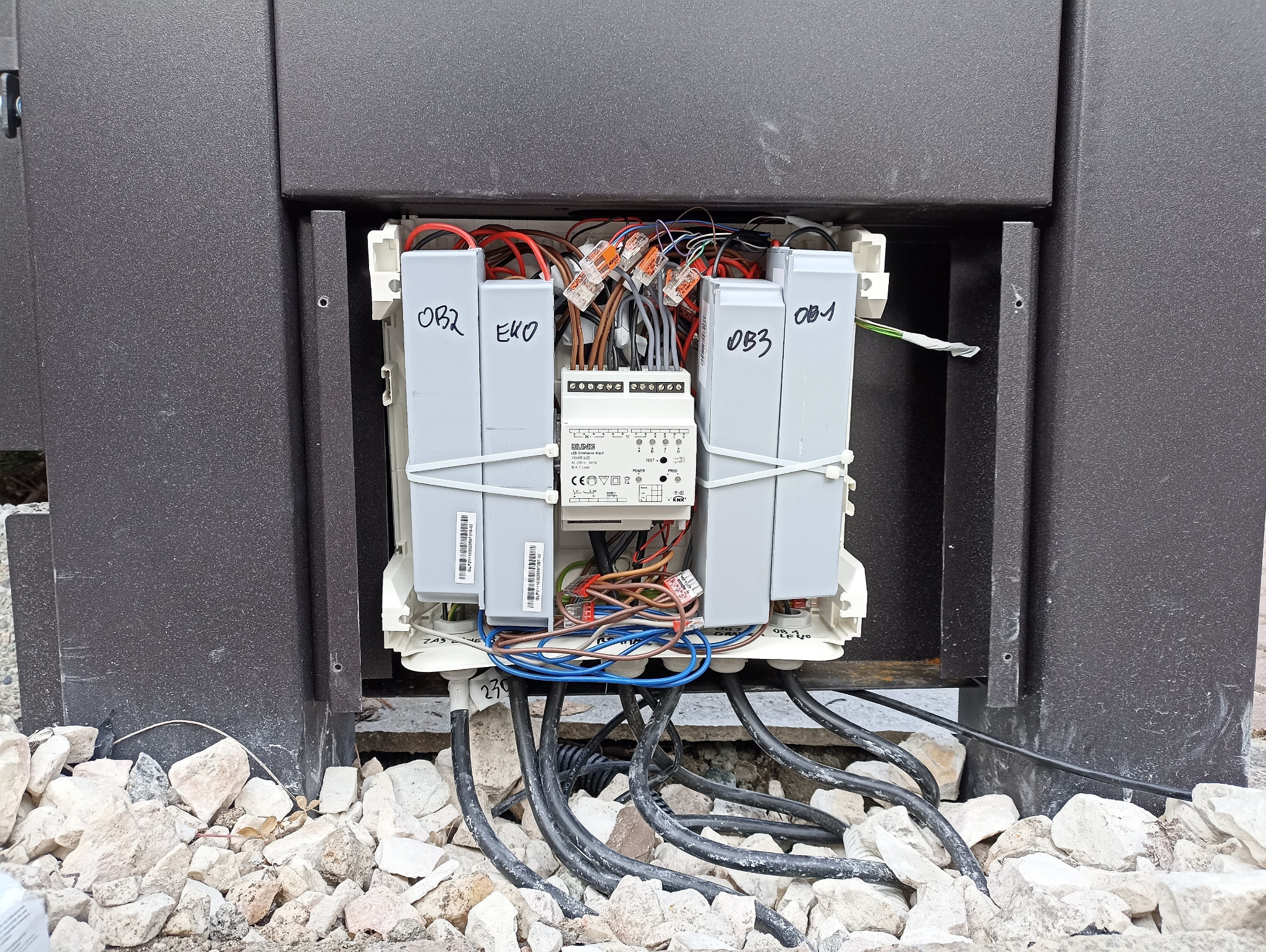 Otwarta skrzynka elektryczna z widocznymi przewodami, złączkami WAGO, modułami z oznaczeniami OB1, OB2, OB3, EKO i centralnym urządzeniem sterującym JUNG, całość umieszczona w ciemnej obudowie...