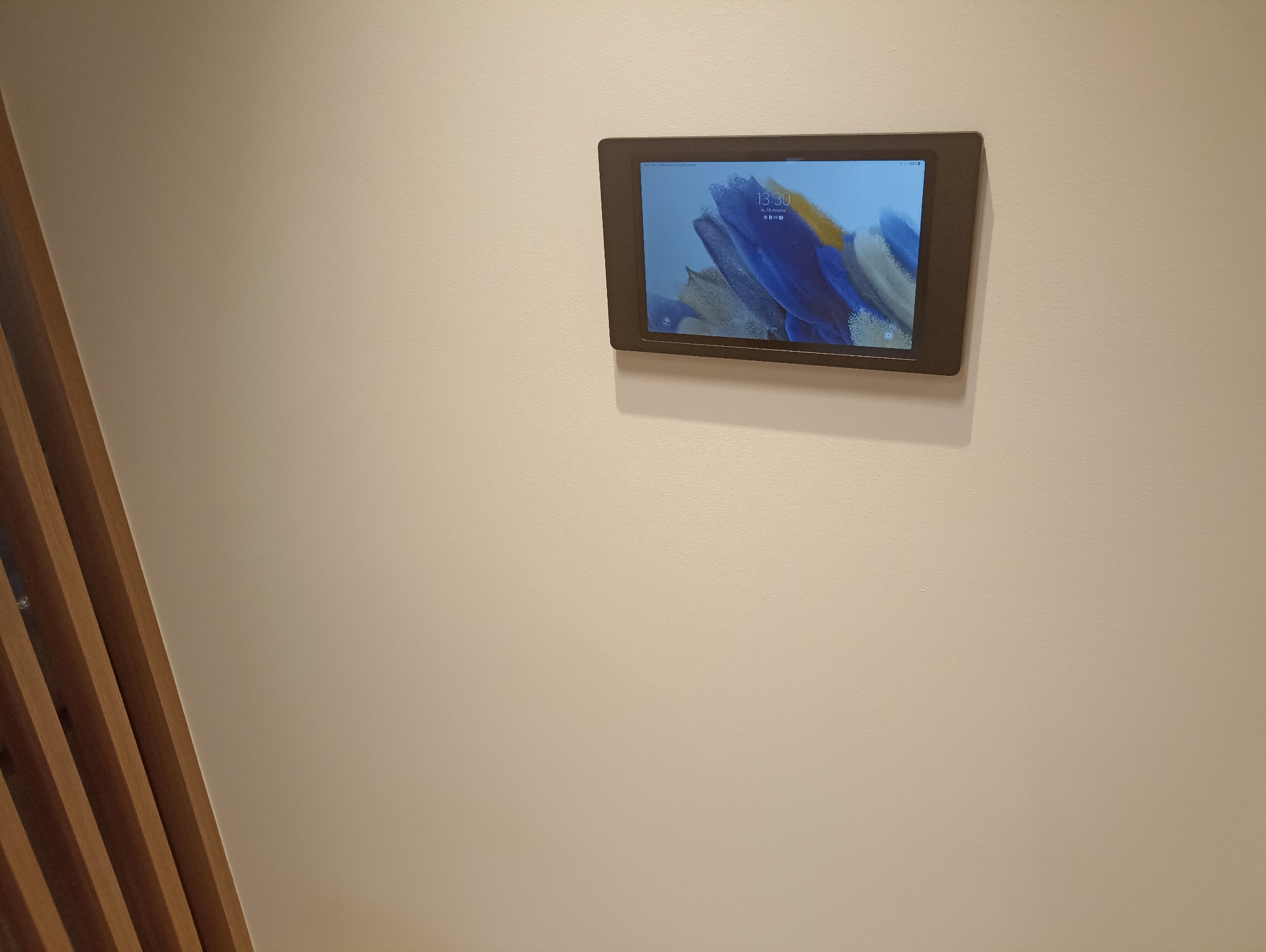Tablet w ciemnej obudowie zamontowany na kremowej ścianie, wyświetlający godzinę 13:30 i datę 11 stycznia, z abstrakcyjną tapetą w odcieniach błękitu i żółci, widoczne drewniane listwy dekoracyjne...