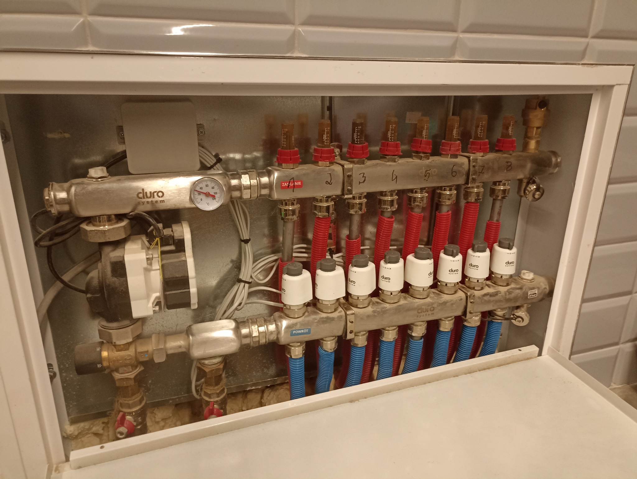 Rozdzielacz ogrzewania podłogowego marki Cluro System zamontowany w szafce, widoczne rury w kolorach czerwonym i niebieskim oraz pompy obiegowe. Instalacja wykonana w miejscowości Sójki.