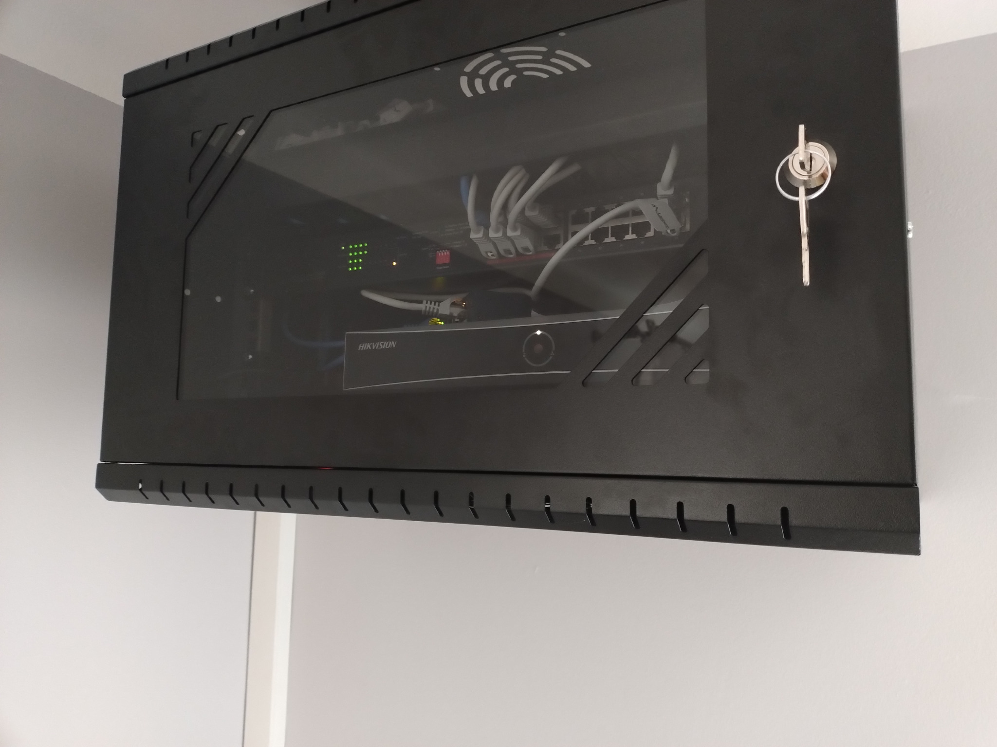 Czarna szafka rack wisząca na ścianie, z widocznym sprzętem sieciowym (router, przełącznik) i okablowaniem. Kluczyk w zamku szafki.
