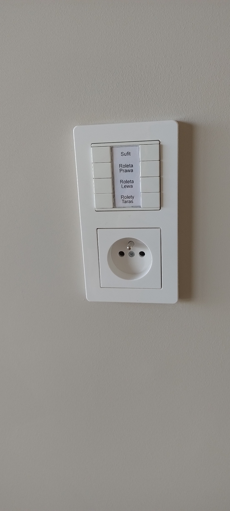 Panel sterowania inteligentnego domu z włącznikami oznaczonymi etykietami: Sufit, Roleta Prawa, Roleta Lewa, Rolety Taras, oraz gniazdkiem elektrycznym, zamontowany na szarej ścianie.