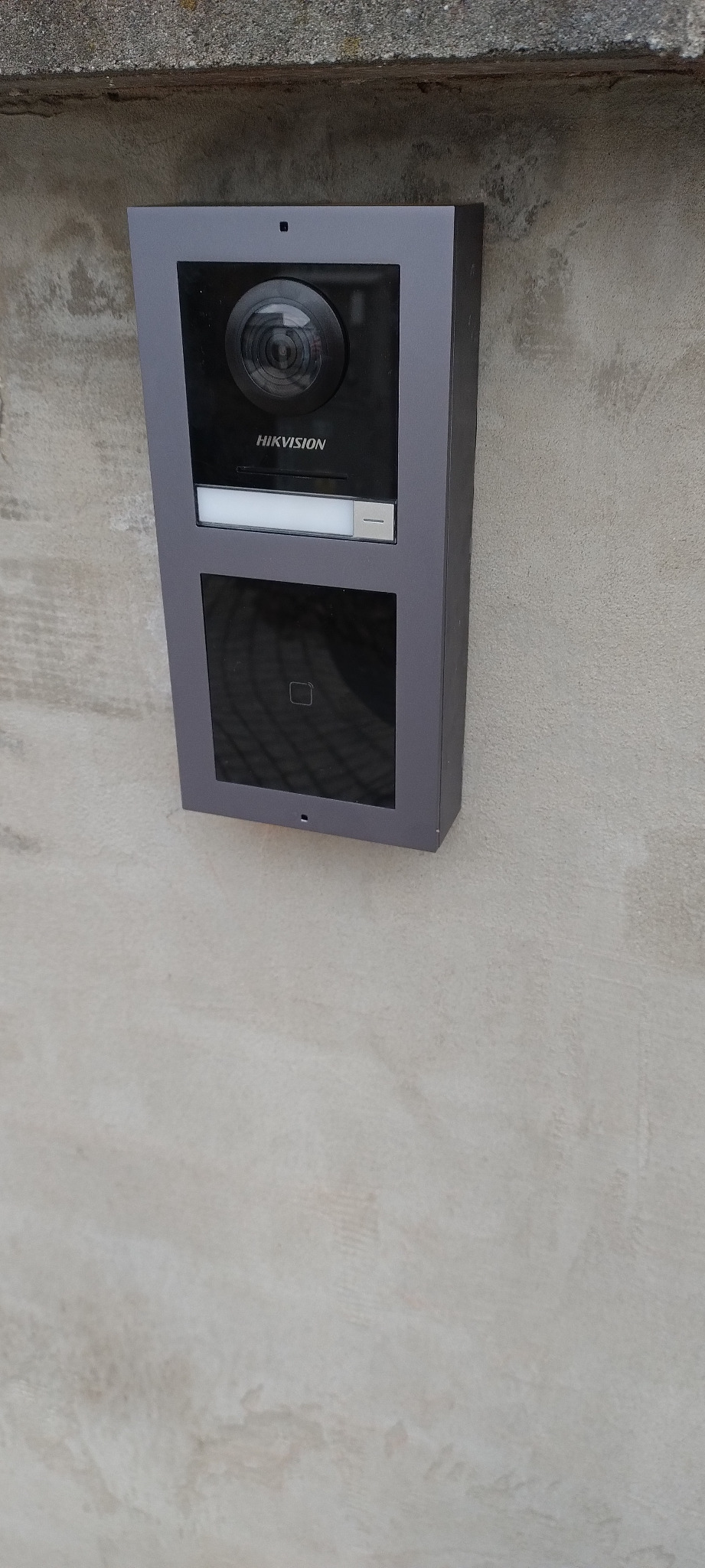 Szary wideodomofon Hikvision zamontowany na jasnej, strukturalnej ścianie zewnętrznej budynku. Widoczna kamera, czytnik zbliżeniowy i podświetlany przycisk.