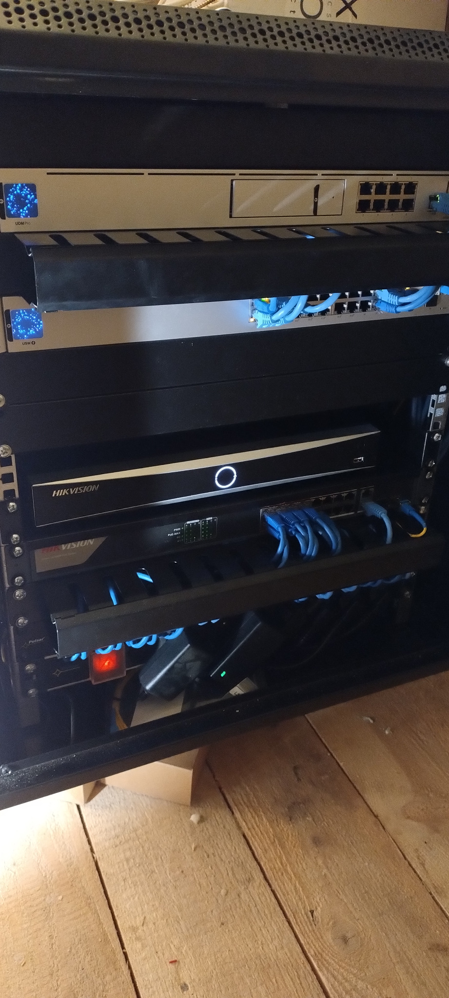 Zamknięta szafa rack z zainstalowanymi urządzeniami sieciowymi, w tym rejestratorem Hikvision, switchami i routerami, połączonymi niebieskimi kablami Ethernet.
