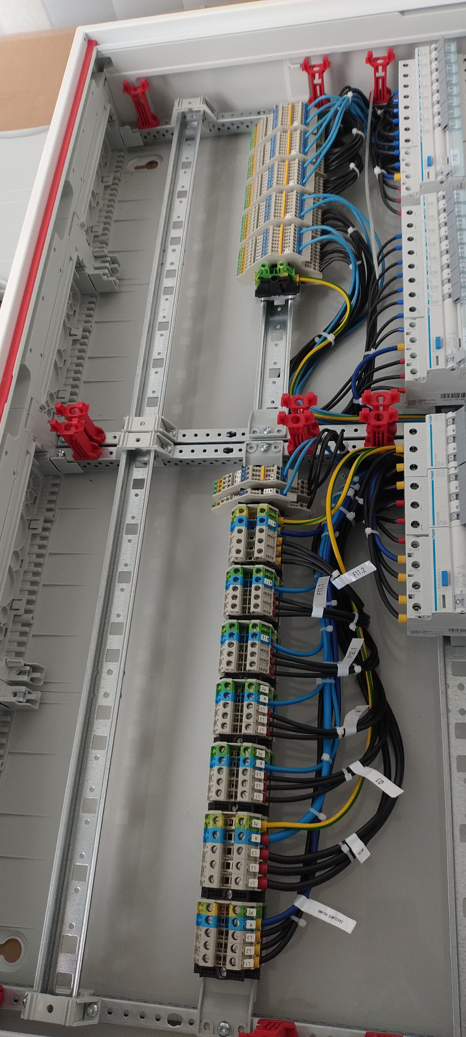 Rozdzielnica elektryczna w trakcie montażu, widoczne kable o różnych kolorach podłączone do listew zaciskowych, oznaczone etykietami. Szyny DIN zamontowane na metalowej konstrukcji wewnątrz obudowy.