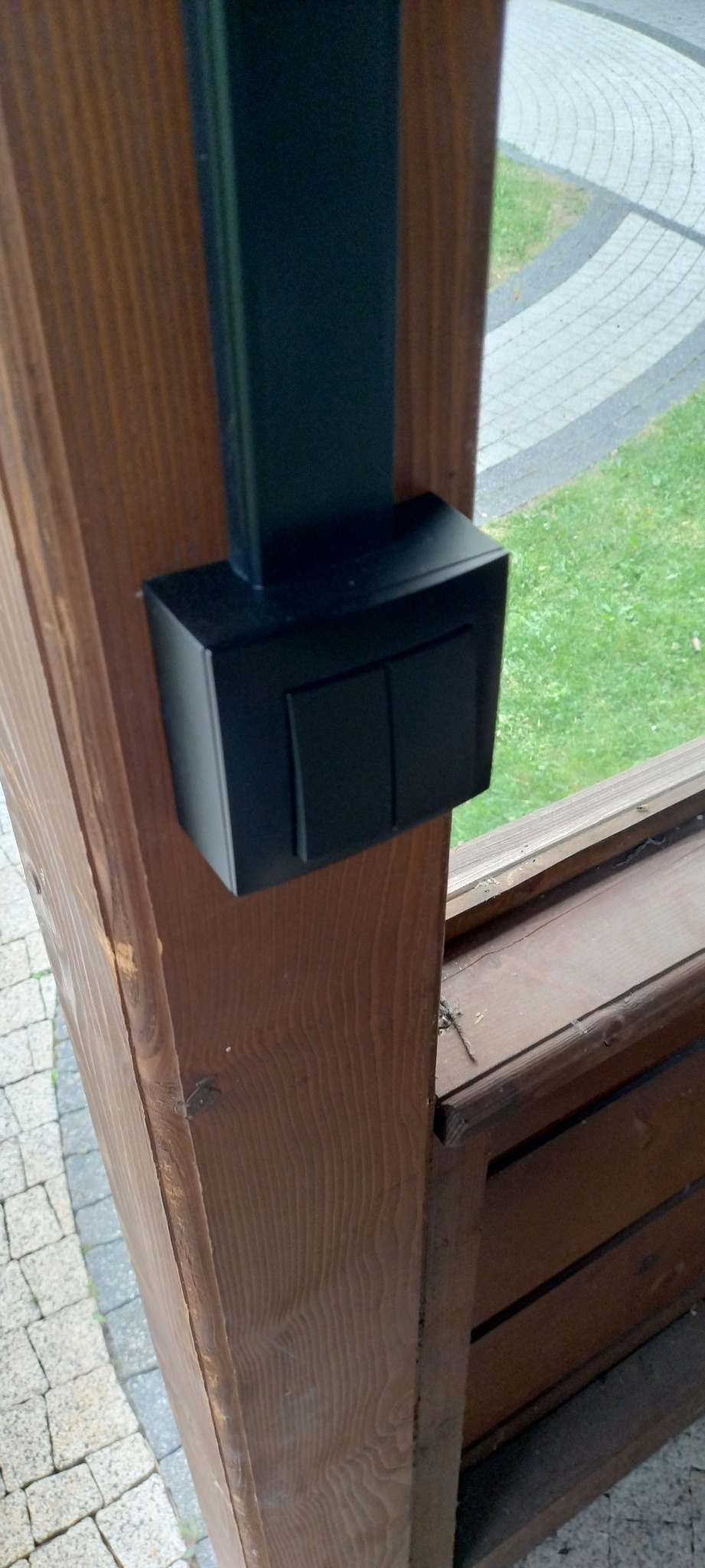 Czarny, podwójny włącznik światła zamontowany na drewnianym słupku altany, widok z bliska, na zewnątrz kostka brukowa i trawnik.