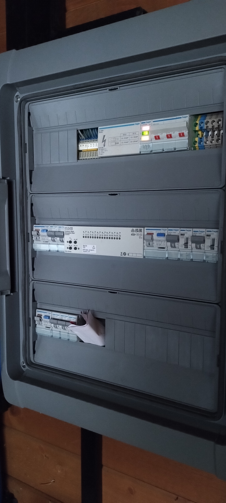 Otwarta skrzynka elektryczna z widocznymi bezpiecznikami, przekaźnikami i modułami sterującymi, w trakcie instalacji lub serwisu, z elementami Hager i Jung.