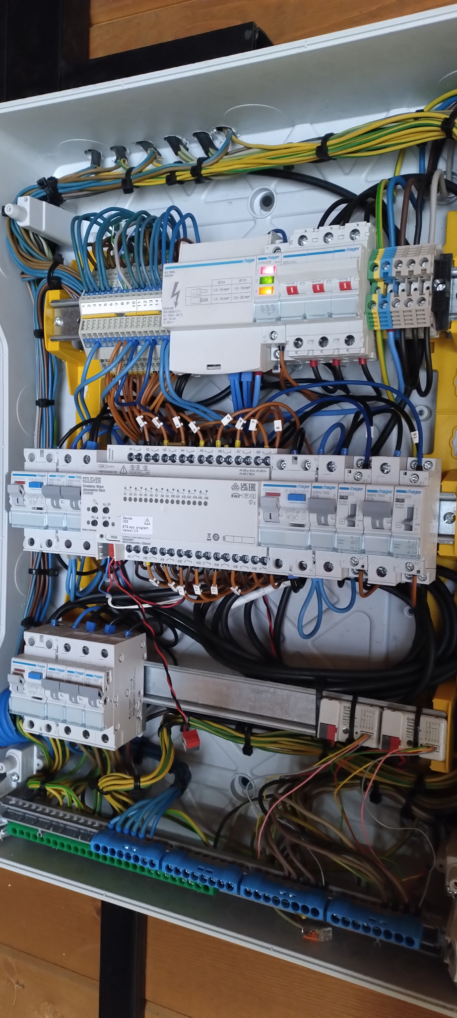 Otwarta skrzynka rozdzielcza z zamontowanymi zabezpieczeniami, modułami sterującymi i licznymi przewodami w kolorach niebieskim, żółto-zielonym i brązowym.