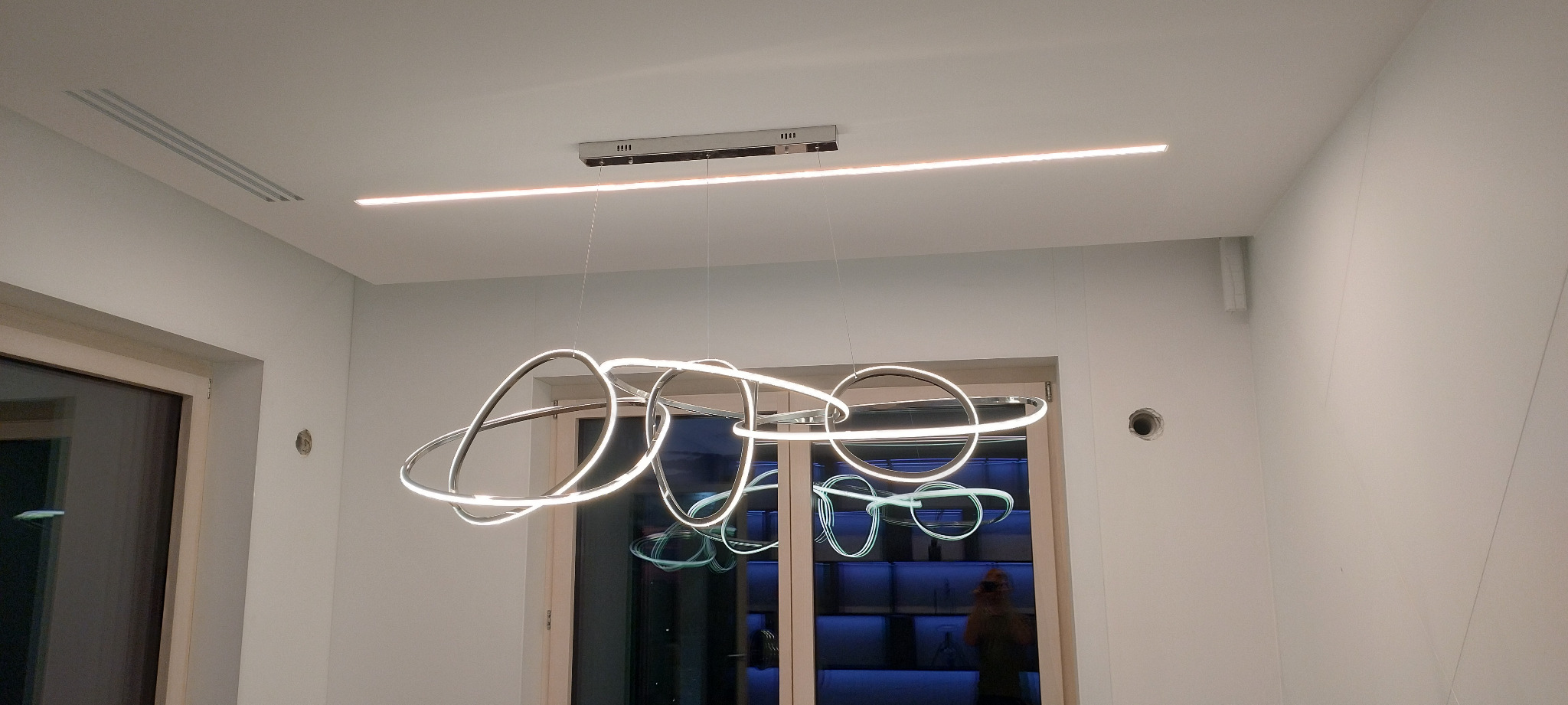 Nowoczesna lampa wisząca z okrągłymi elementami LED i listwa oświetleniowa LED na suficie w minimalistycznym wnętrzu z widoczną wentylacją sufitową i otworem na ścianie.