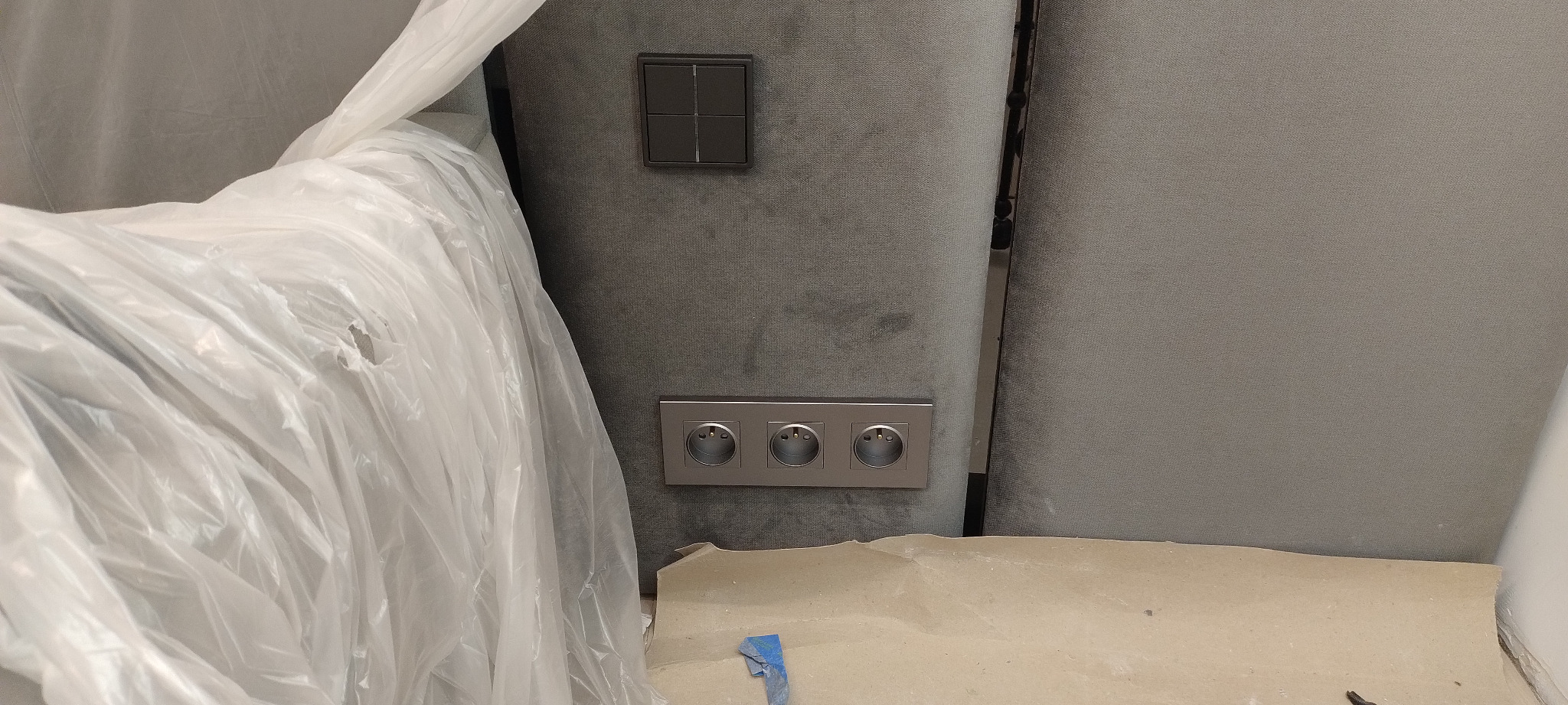 Szare gniazdka elektryczne i włącznik światła na ścianie, częściowo zasłonięte folią ochronną i papierem malarskim.