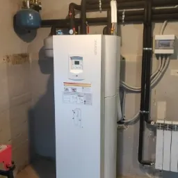 Biała pompa ciepła typu split firmy Atlantic zainstalowana w pomieszczeniu technicznym, widoczne rury, zbiornik wyrównawczy i skrzynka elektryczna.