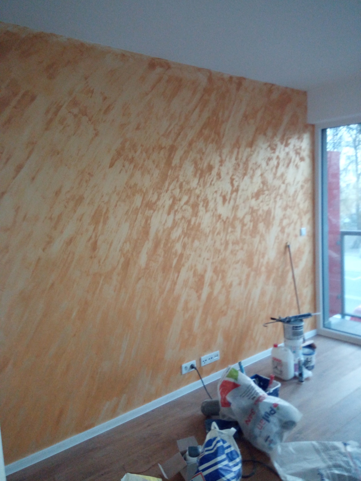 Ściana w odcieniach beżu i brązu z dekoracyjnym efektem malarskim, widoczne gniazdka elektryczne, podłoga z paneli i akcesoria malarskie w rogu pokoju.