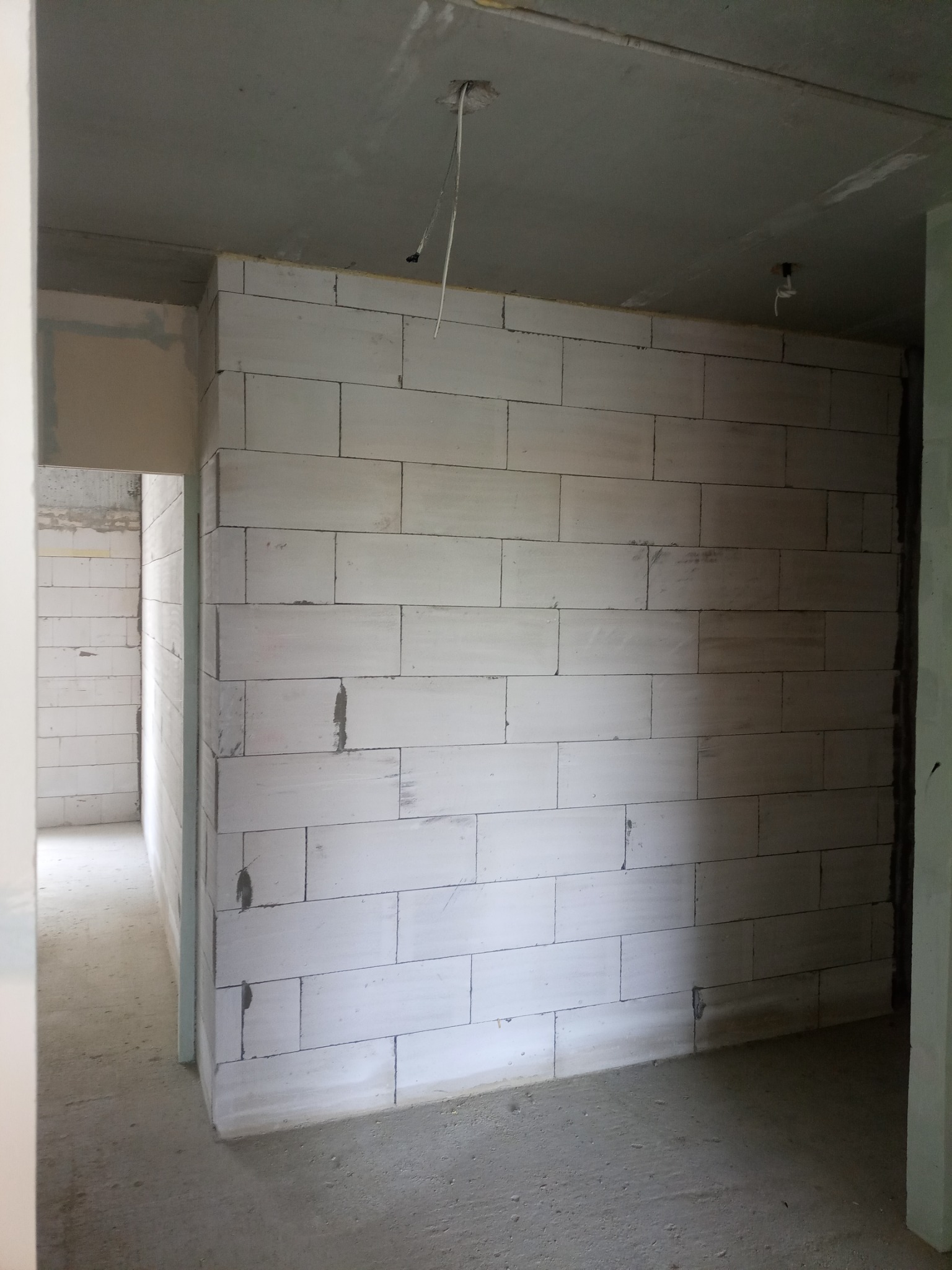 Świeżo wymurowana ściana z białych bloczków w surowym stanie, widoczne przewody elektryczne wystające z sufitu, szara podłoga i fragmenty innych ścian w tle.