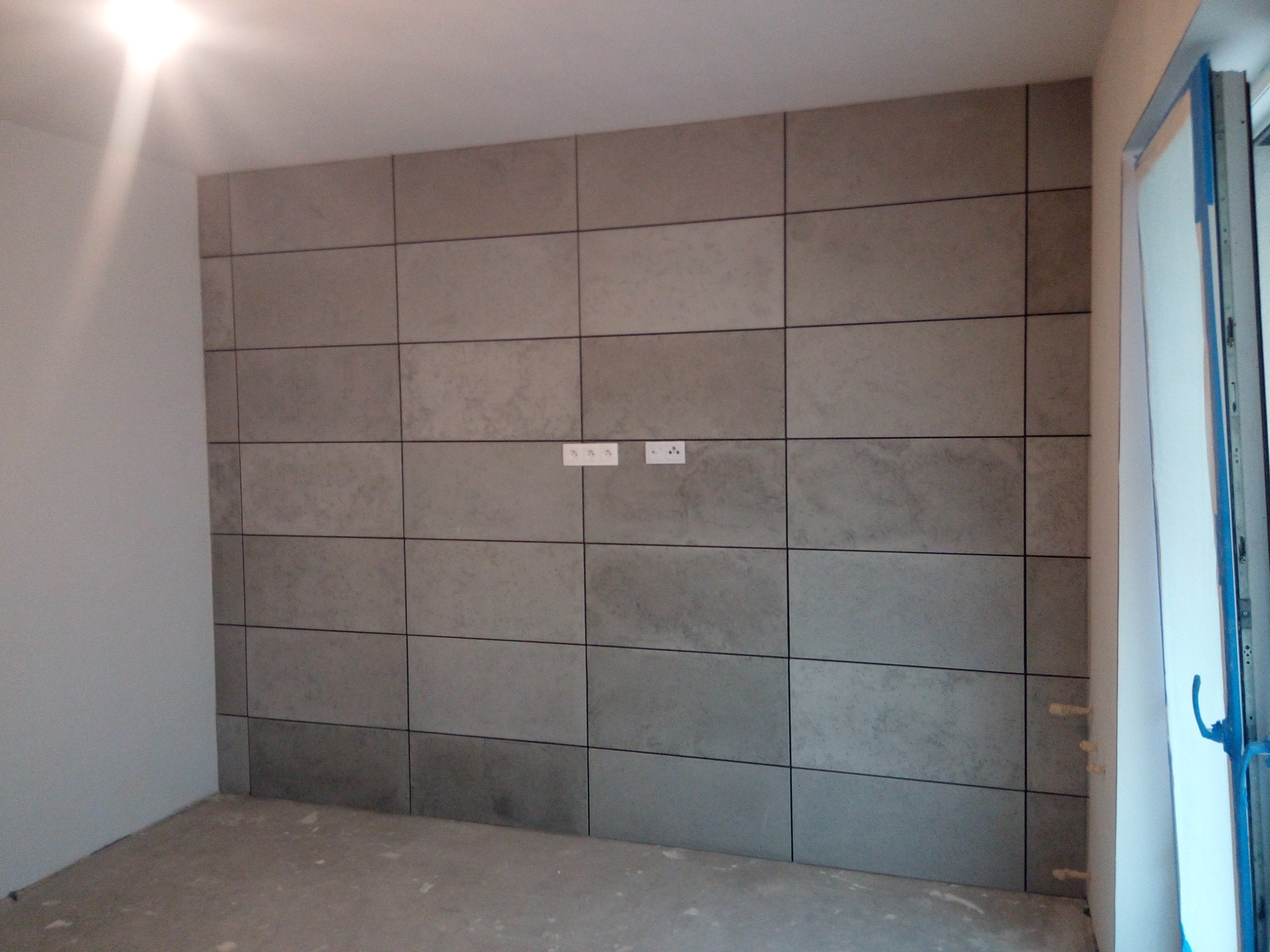 Ściana wykończona płytami w odcieniach szarości z widocznymi fugami, gniazdka elektryczne na środku, surowa podłoga i fragment ościeżnicy drzwi.