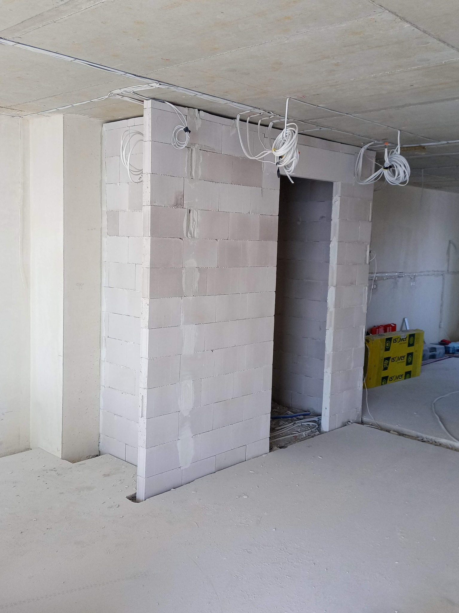 Wnętrze w stanie surowym: wymurowana ściana z pustaków z otworem drzwiowym, widoczne przewody elektryczne zwisające z sufitu, surowy beton na podłodze i ścianach, materiały izolacyjne w tle.