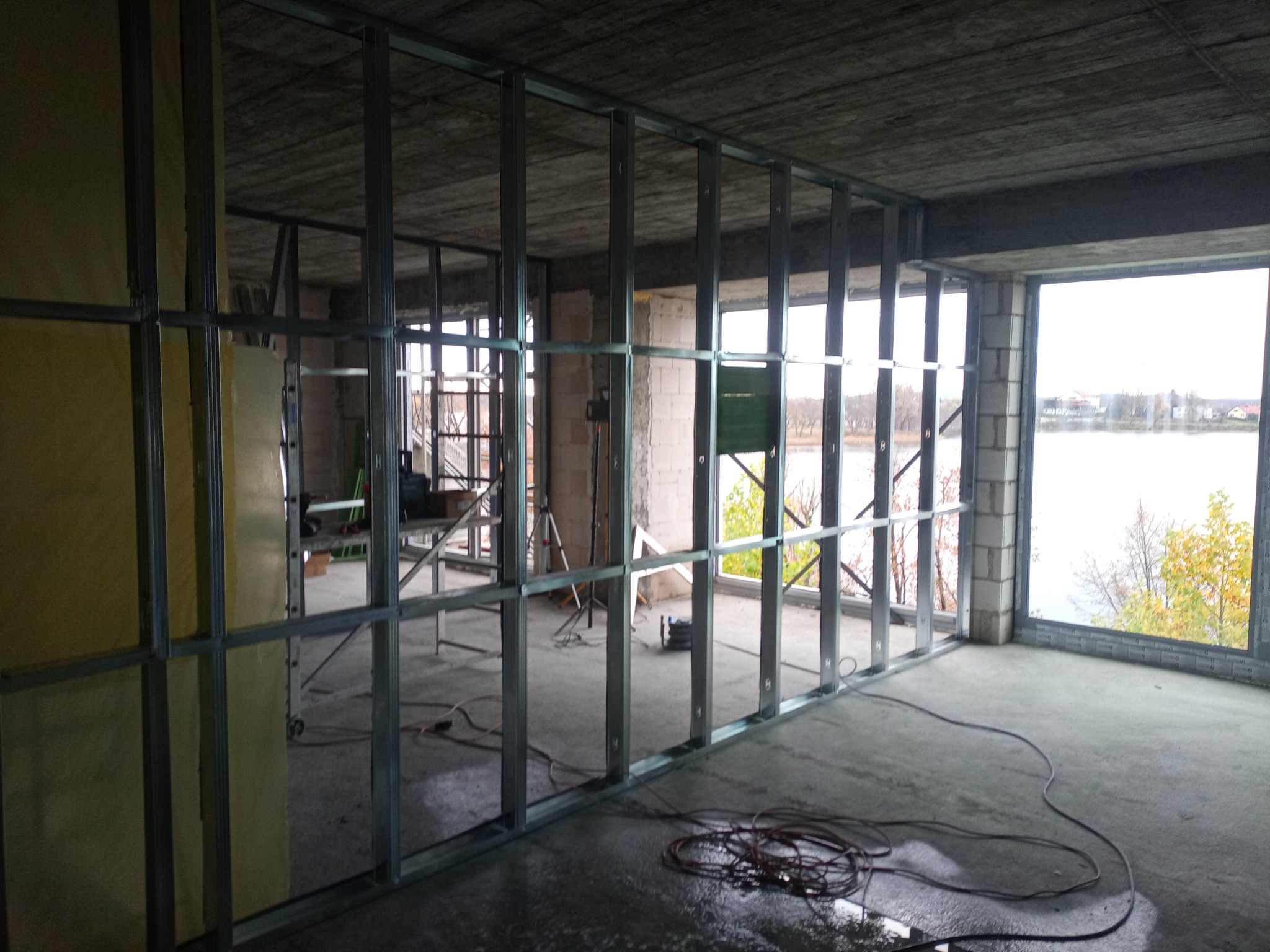 Konstrukcja szkieletowa z profili stalowych, widok na surowy betonowy strop i okno z widokiem na jezioro, przewody elektryczne na mokrej posadzce.
