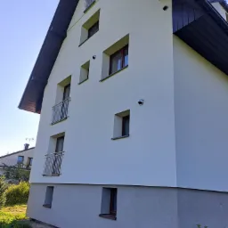 Nowoczesny dom jednorodzinny z jasną elewacją i ciemnym dachem, widoczne małe balkony z metalowymi barierkami, ocieplenie dwoma kolorami tynku, widoczne otwory wentylacyjne na elewacji.