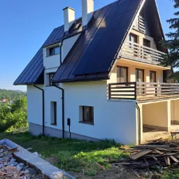 Nowoczesny dom z ciemnym dachem i jasną elewacją, widok z zewnątrz na tle zieleni i błękitnego nieba, z widocznymi pozostałościami po budowie w otoczeniu.