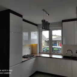 Nowoczesna kuchnia z białymi frontami szafek, ciemnobrązowymi bokami i drewnianym blatem, oświetlona lampą wiszącą z trzema cylindrycznymi kloszami, widok z okna na zabudowania i samochód.