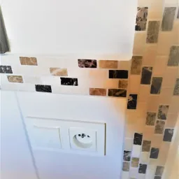 Fragment ściany z mozaiką w odcieniach beżu i brązu, ułożoną w pasy, obok gniazdka elektrycznego z włącznikiem w białej ramce.